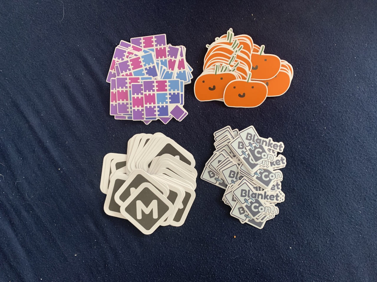 Four piles of stickers arranged in a 2x2 grid. From left to right: Quilt logo, Pineapple, Modfest logo, BlanketCon 2022 logo